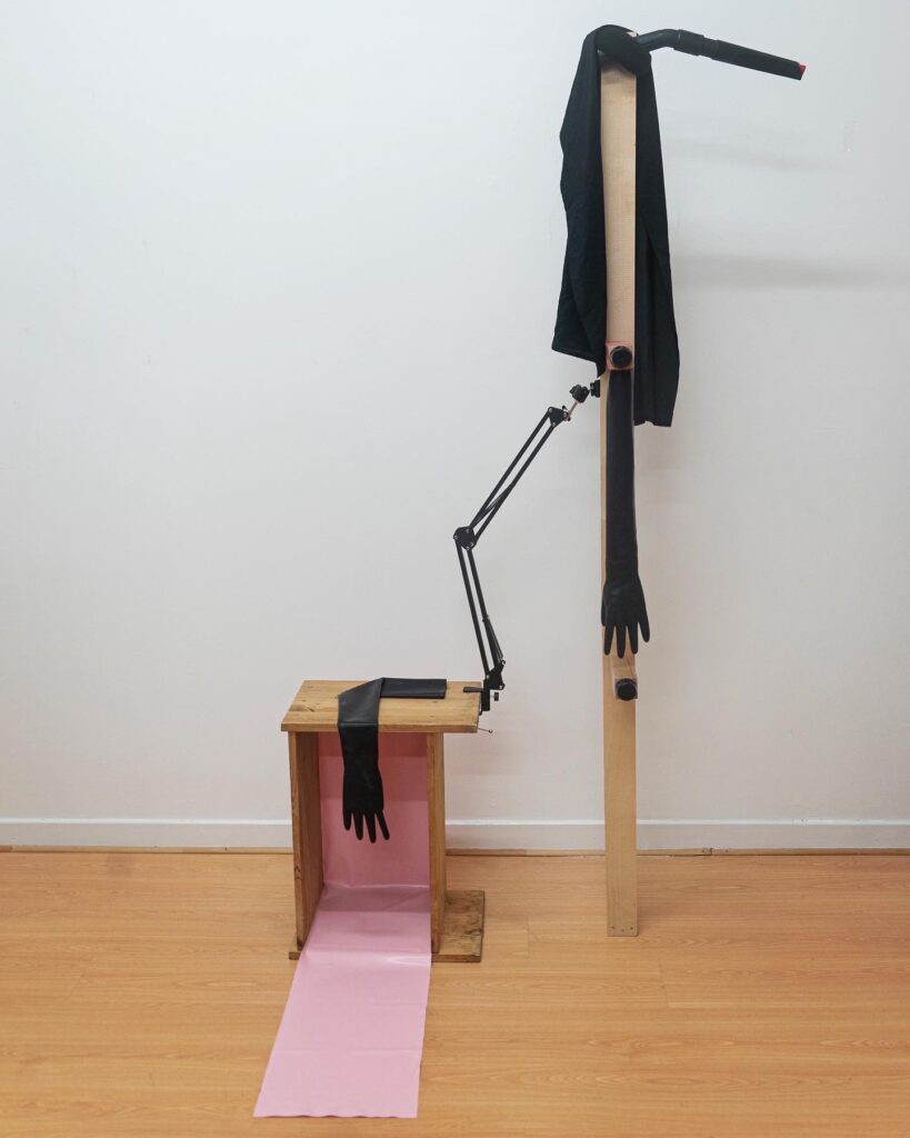 Sculptural installation with a tall wooden structure, black fabric arms and a pale pink floor-length material